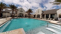 This expansive outdoor amenity space showcases a resort-style design that seamlessly blends architectural elegance with leisure-focused functionality. At its heart lies a large, crystal-clear swimming pool with vivid turquoise water, surrounded by a generously sized travertine-style pool deck. A standout feature is the in-pool sun shelf with built-in lounge chairs and water jets, offering a relaxing wading experience just inches below the surface.

The centerpiece of the space is a Spanish Colonial-inspired clubhouse, distinguished by its crisp white stucco facade, dramatic stepped parapet gables, tall arched windows, and classic wall-mounted lanterns. The building's architecture lends a sophisticated, upscale character to the entire outdoor environment.

Towering palm trees are thoughtfully positioned throughout the pool deck, providing natural shade and reinforcing the tropical, resort-like atmosphere. Rows of cushioned chaise lounges and poolside chairs are arranged along the deck, offering ample space for sunbathing and relaxation. Shade umbrellas are stationed in select areas, providing additional comfort on sunny days.

The surrounding multi-story residential buildings, featuring balconies and large windows, frame the courtyard and overlook the pool area. Manicured landscaping with tropical plantings adds lush greenery throughout, enhancing the overall ambiance of this well-appointed outdoor retreat.