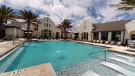 This expansive outdoor amenity space showcases a resort-style design that seamlessly blends architectural elegance with leisure-focused functionality. At its heart lies a large, crystal-clear swimming pool with vivid turquoise water, surrounded by a generously sized travertine-style pool deck. A standout feature is the in-pool sun shelf with built-in lounge chairs and water jets, offering a relaxing wading experience just inches below the surface.

The centerpiece of the space is a Spanish Colonial-inspired clubhouse, distinguished by its crisp white stucco facade, dramatic stepped parapet gables, tall arched windows, and classic wall-mounted lanterns. The building's architecture lends a sophisticated, upscale character to the entire outdoor environment.

Towering palm trees are thoughtfully positioned throughout the pool deck, providing natural shade and reinforcing the tropical, resort-like atmosphere. Rows of cushioned chaise lounges and poolside chairs are arranged along the deck, offering ample space for sunbathing and relaxation. Shade umbrellas are stationed in select areas, providing additional comfort on sunny days.

The surrounding multi-story residential buildings, featuring balconies and large windows, frame the courtyard and overlook the pool area. Manicured landscaping with tropical plantings adds lush greenery throughout, enhancing the overall ambiance of this well-appointed outdoor retreat.