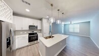 This thoughtfully designed single-floor residence offers a clean, modern aesthetic throughout, combining functionality with contemporary style. The open-concept kitchen is a standout feature, anchored by a large center island with a white quartz countertop and an undermount sink, complemented by elegant pendant lighting with brass chain accents. Crisp white shaker cabinetry, grey subway tile backsplash, and a full suite of stainless steel appliances — including a French door refrigerator, electric range, microwave, and dishwasher — complete the kitchen's polished look. Light wood-tone luxury vinyl plank flooring flows seamlessly from the kitchen into the living area, which is bathed in natural light from multiple windows and features a ceiling fan overhead. A glass door provides additional light and access to the exterior. The bedroom offers a comfortable retreat with plush carpeting, a ceiling fan, and a bright window with blinds. The bathroom is finished with a white vanity, bold-framed mirror, and a glass-enclosed tub-shower combination with marble-style tile surround. A dedicated laundry room includes a full-size washer and dryer alongside built-in shelving and a closet rod, adding exceptional convenience to everyday living.