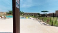 This outdoor recreational area centers around two distinct pools: a generously sized main swimming pool with a classic kidney-shaped design, and a separate circular wading pool, both finished with brick-edged borders and surrounded by a spacious concrete deck. The clear, turquoise water in both pools reflects the open sky, creating a bright and inviting atmosphere. The deck is furnished with multiple suntanning lounge chairs, providing comfortable spots for relaxation throughout the day. A covered pergola with a green shade canopy offers a sheltered retreat from direct sunlight, making the space functional even during peak afternoon hours. The entire pool area is enclosed by a black iron security fence, ensuring safety and privacy. Well-kept green lawns border the perimeter of the fenced zone, adding a natural, refreshing contrast to the hardscape. In the background, the surrounding brick buildings frame the space, giving the area a cohesive and established community feel. Mature trees are visible beyond the fence line, contributing to a pleasant outdoor ambiance. Signage throughout the area enforces safety rules, including no-diving and no-smoking policies, reflecting a commitment to a safe and enjoyable environment for all users.