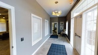 This single-floor space makes an immediate impression with its well-appointed entryway, setting a refined tone throughout. The foyer features large-format tile flooring, crisp white baseboards, and soft neutral grey walls that create a clean, contemporary aesthetic. A modern sputnik-style chandelier hangs overhead, providing a stylish focal point and warm ambient lighting to greet visitors upon entry. Large windows with white-trimmed blinds flank the entrance, allowing natural light to filter in and brighten the space. The entryway transitions seamlessly into an adjacent sitting area, visible through a framed doorway, where textured dark accent wallpaper adds depth and visual interest. A cozy armchair and side table are positioned beneath framed artwork, creating an intimate nook that balances style and function. The overall design palette — combining warm greys, crisp whites, and dark accents — gives the space a cohesive, sophisticated character. The layout is thoughtfully arranged, offering a smooth flow from the entry into the interior, making the space feel both functional and welcoming from the moment you step inside.