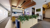 Located at 4557 N Sheridan Road in Chicago's Uptown neighborhood, this single-floor lobby and common area space showcases a thoughtfully designed, contemporary interior. The welcoming entrance features floor-to-ceiling glass walls and doors that flood the space with natural light and offer clear views of the surrounding streetscape. A distinctive slatted wood ceiling adds warmth and architectural interest, creating a refined contrast against the crisp white and charcoal-toned walls throughout. The main lounge area is furnished with a tufted navy velvet sofa, sculptural lounge chairs, and a round marble-top coffee table, all anchored by a textured area rug. Lush, oversized planters filled with leafy greenery serve as natural dividers, enhancing the biophilic character of the space. A living moss wall art piece adds an organic, decorative accent. The wet bar area features sleek cabinetry, a marble backsplash, and integrated appliances, complemented by mounted flat-screen displays. Glass-partitioned private offices provide semi-transparent workspaces with ample natural light. A mailbox alcove, parcel locker, and elevator access are conveniently located along a well-appointed corridor. The overall ambiance is polished and professional, balancing functionality with an inviting, upscale aesthetic.