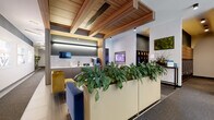 Located at 4557 N Sheridan Road in Chicago's Uptown neighborhood, this single-floor lobby and common area space showcases a thoughtfully designed, contemporary interior. The welcoming entrance features floor-to-ceiling glass walls and doors that flood the space with natural light and offer clear views of the surrounding streetscape. A distinctive slatted wood ceiling adds warmth and architectural interest, creating a refined contrast against the crisp white and charcoal-toned walls throughout. The main lounge area is furnished with a tufted navy velvet sofa, sculptural lounge chairs, and a round marble-top coffee table, all anchored by a textured area rug. Lush, oversized planters filled with leafy greenery serve as natural dividers, enhancing the biophilic character of the space. A living moss wall art piece adds an organic, decorative accent. The wet bar area features sleek cabinetry, a marble backsplash, and integrated appliances, complemented by mounted flat-screen displays. Glass-partitioned private offices provide semi-transparent workspaces with ample natural light. A mailbox alcove, parcel locker, and elevator access are conveniently located along a well-appointed corridor. The overall ambiance is polished and professional, balancing functionality with an inviting, upscale aesthetic.