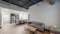 Located at 2611 E Third St in Bloomington, Indiana, this three-bedroom, three-bathroom unit offers a modern industrial aesthetic across a single floor. The open-concept living and kitchen area immediately impresses with soaring exposed ceilings featuring dark metal beams and ductwork, creating a loft-like atmosphere. Wood-look plank flooring runs throughout the common areas, adding warmth to the contemporary design. The living room provides ample space for entertaining, with a large window allowing natural light to fill the room. The kitchen is well-appointed with stainless steel appliances, including a refrigerator, gas range, and microwave, complemented by dark cabinetry and a bright white countertop island with bar seating — ideal for casual dining. Two hallways connect the living spaces to the three bedrooms, each featuring carpeted flooring and a dedicated window. The primary bedroom offers generous proportions, while the additional bedrooms are well-sized and functional. Each of the three bathrooms is cleanly finished with light flooring, dark vanity cabinetry, and white countertops. The unit also includes a dedicated laundry area and multiple closets, providing practical storage solutions throughout.
