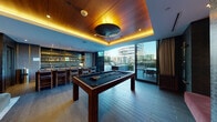 Located at 1755 Argyle Ave in the heart of Hollywood, Argyle House is a thoughtfully designed amenity space that seamlessly blends indoor comfort with outdoor living. The interior lounge is anchored by a striking billiards table set beneath a warm wood-paneled tray ceiling with ambient cove lighting, creating an inviting atmosphere. A full bar area features a layered stone backsplash, under-cabinet illumination, open shelving, and generous counter seating with upholstered bar stools — ideal for social gatherings. The lounge area is furnished with plush seating and curated artwork, adding a refined, gallery-like quality to the space. Dark textured wall treatments and tile flooring throughout contribute to the space's moody, upscale aesthetic. Floor-to-ceiling sliding glass doors dissolve the boundary between indoors and out, opening directly to a spacious stone-tiled terrace. The outdoor area features a fully equipped BBQ and prep station, manicured planters, and alfresco dining, all set against a striking backdrop — the iconic Capitol Records Building rising prominently in the skyline. The entry foyer features richly textured dark walls, curated furnishings, and a decorative mirror, setting an immediate tone of sophistication upon arrival.