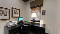 This business center is a focused, efficiently designed workspace intended to meet the productivity needs of guests. The room is anchored by a dark wood L-shaped desk that provides ample surface area for working, accommodating a desktop computer with monitor, a keyboard, and a mouse. A printer is conveniently positioned at one end of the desk, offering easy access for printing documents. A sleek table lamp provides warm, localized lighting to complement the room's ambient illumination, creating a comfortable environment for extended use. A framed piece of artwork adorns one wall, adding a touch of polish to the otherwise utilitarian space. Natural light filters in through a window dressed with horizontal blinds and a decorative valance, contributing to a pleasant and focused atmosphere. Posted computer use rules and a clearly visible no-smoking sign reflect the space's commitment to an orderly and respectful environment for all users. The neutral wall tones and dark furnishings lend the room a professional character. Overall, this business center offers a quiet, well-equipped setting for guests who need to stay connected, print materials, or attend to work tasks during their stay.