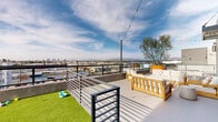 Located at 7650 Van Nuys Blvd in Los Angeles, this rooftop amenity deck offers a well-appointed outdoor retreat designed for both relaxation and pet-friendly recreation. The space is defined by its modern architectural backdrop — smooth concrete facades, large glass doors, and sleek metal railings — all set against an expansive open sky with sweeping panoramic views of the San Fernando Valley and the surrounding mountain ranges.

The deck is divided into two distinct functional zones. The lounge area features a generous seating arrangement of solid wood-framed sofas and chairs with plush white cushions and vibrant patterned accent pillows, centered around a matching wood coffee table. String lights are strung overhead, adding a warm ambiance for evening use. Large windows and glass doors connect the deck to the interior, allowing natural light to flow freely between the spaces.

The second zone is a dedicated pet area, enclosed by a metal railing and surfaced with bright artificial turf. Pet amenities including water bowls and toys are provided, making it a practical and engaging space for animals. Planters with ornamental trees add greenery and visual warmth throughout the deck. Together, these elements create a versatile, community-oriented rooftop space that balances comfort, utility, and striking elevated views of the greater Los Angeles area.