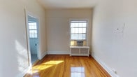 This single-floor space showcases a clean, classic interior defined by warm hardwood floors that catch and reflect natural sunlight beautifully. The main room features tall walls with clean white trim and baseboards, contributing to an airy, open feel. A multi-pane double-hung window allows generous natural light to fill the space while offering exterior views, and a window-mounted air conditioning unit ensures comfortable cooling during warmer months. A white radiator beneath the window provides supplemental heating, suggesting a traditional heating system typical of well-established residential buildings. The neutral, light beige wall tones create a versatile backdrop suitable for various interior styles. A doorway visible from the main room leads to an adjacent space, hinting at a functional layout with connected areas. The overall ambiance is bright, understated, and practical, with classic architectural elements that reflect a timeless residential character. The well-maintained hardwood floors and consistent trim work throughout suggest careful upkeep, making this a move-in ready space with lasting structural appeal.