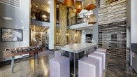 This dynamic communal lounge showcases an impressive industrial-modern aesthetic with soaring double-height ceilings that lend a grand sense of openness to the space. At the heart of the main floor sits a substantial communal table with a concrete-style surface, surrounded by plush upholstered stools, creating an inviting hub for gathering and collaboration. Overhead, a striking cluster of geometric copper and metallic pendant lights serves as a dramatic focal point, casting warm ambient light throughout the room. Textured vertical stone or wood-clad columns anchor the space, adding natural warmth and visual depth against the polished concrete floors and neutral-toned walls. A mezzanine level, accessible by the visible upper balcony with sleek metal railings, overlooks the main lounge area, enhancing the vertical drama of the design. To one side, a foosball table signals the space's recreational character, while display shelving and curated décor elements contribute to the overall sophisticated, urban atmosphere. Slatted wooden panels and industrial-style metal accents throughout further reinforce the cohesive modern-industrial design language, making this a well-appointed and multifunctional social environment.