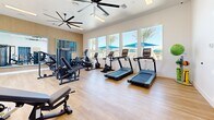 Located in Phoenix, AZ, this dedicated fitness center offers a thoughtfully designed exercise space spread across a single level. The room features light hardwood-style flooring throughout, lending a clean and modern aesthetic that complements the bright, open atmosphere.

Large windows line one wall, flooding the space with abundant natural light and offering views of palm trees and vibrant blue poolside umbrellas, creating an energizing workout environment. Dual large ceiling fans overhead ensure air circulation throughout the room.

The fitness center is well-appointed with a comprehensive selection of equipment, including multiple treadmills, an elliptical trainer, a stationary bike, a multi-functional cable and strength machine, a dedicated bench press station, and a full dumbbell rack — accommodating a wide range of workout routines. A wall-mounted flat-screen television adds entertainment value for cardio sessions.

A full-length framed mirror on a wood-paneled accent wall enhances the sense of space. A built-in water fountain station with a bold blue geometric tile surround provides a functional and stylish hydration point. The entry features sleek black-framed glass double doors, with an adjacent open area that could serve as a stretching or warm-up zone. The overall design balances functionality with a polished, contemporary finish ideal for an active community.