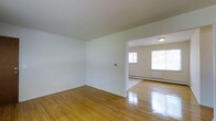 This single-floor space presents a clean, straightforward layout with an emphasis on simplicity and functionality. The interior is defined by warm, polished hardwood floors that run throughout, reflecting natural light and adding a timeless character to the space. Crisp white walls provide a bright, neutral backdrop that enhances the sense of openness across each room. The main living area flows seamlessly into an adjacent room through a wide, open doorway, creating a connected and airy feel. The adjoining room benefits from natural light streaming in through a large window, further brightening the interior and offering views of the surrounding greenery. A baseboard heating unit lines the wall of the secondary room, ensuring functional climate control. Flush-mount ceiling lighting provides clean, even illumination throughout. The overall ambiance is understated and move-in ready, offering a versatile canvas suited to a range of furnishing styles. The straightforward, single-level design makes the space easy to navigate, while the consistent flooring and neutral tones create a cohesive and harmonious environment throughout.