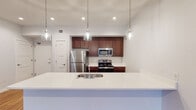 Located on E Market Street in the heart of Greensboro, NC, this single-floor unit offers a thoughtfully designed open-concept layout with contemporary finishes throughout. The kitchen serves as a central feature, equipped with rich dark-wood cabinetry, stainless steel appliances including a refrigerator, microwave, and range, and a herringbone tile backsplash. A large white quartz peninsula with an undermount sink and elegant pendant lighting overhead provides both ample workspace and a natural gathering point that flows seamlessly into the living area. Warm hardwood-style flooring extends throughout the main living spaces, complementing the clean white walls and recessed lighting that create a bright, airy atmosphere. The spacious living area benefits from a wall of floor-to-ceiling sliding glass doors leading to a private balcony, filling the space with natural light and outdoor views. The bedrooms feature large modern windows with views of the surrounding landscape and ceiling fans for added comfort. The bathrooms are finished with marble-style tile flooring, rich wood vanities with white countertops, and well-lit mirrors. One bathroom includes a glass-enclosed walk-in shower, while the second serves as a full bath. The overall design balances modern urban aesthetics with warm, residential comfort.