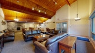 This single-floor communal space is defined by its striking architectural character, most notably the soaring vaulted ceiling clad in warm natural wood planks and supported by exposed wooden beams. Track lighting runs along the beams, illuminating the expansive open interior and enhancing its lodge-like ambiance.

The main gathering area is generously appointed with multiple large leather sofas and armchairs arranged around wooden coffee tables, creating comfortable lounging zones ideal for group gatherings. A wall-mounted flat-screen television and media console provide an entertainment focal point in one seating nook, complemented by natural light from nearby windows.

The dining area features two large wooden tables, each surrounded by cushioned wood chairs, offering ample seating for group meals. A standout architectural detail is the arched brick fireplace, which serves as a dramatic focal point and adds warmth and rustic charm to the space.

The fully equipped kitchen is outfitted with light maple cabinetry, granite countertops, stainless steel appliances including a refrigerator, range with hood vent, and microwave, along with a spacious island with bar seating for additional casual dining. Hardwood flooring grounds the kitchen area, while plush carpet covers the main gathering zones, contributing to the overall cozy, welcoming character of this well-appointed communal space.