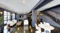 This contemporary space showcases a refined industrial aesthetic, blending modern design elements with functional versatility. The open-plan layout is anchored by striking polished concrete floors that reflect light and add depth to the environment. Exposed metallic ductwork runs along the ceiling, complementing the space's urban character, while recessed lighting ensures a well-lit atmosphere throughout.

The main area is thoughtfully arranged with distinct zones for lounging and meeting. A comfortable seating arrangement features a white sofa and matching accent chairs grouped around a rustic wooden coffee table, creating an inviting conversational space. Nearby, a round marble-top table paired with sleek white modern chairs offers an ideal spot for informal gatherings or collaborative work.

A built-in entertainment wall with a flat-screen display serves as a focal point, framed by bold, dark paneled walls that add a sense of sophistication and contrast against the lighter furnishings. A refreshment station is visible in the background, adding convenience and a welcoming touch. Pops of greenery from potted plants introduce a natural element, softening the industrial feel. Large windows allow natural light to filter in, further enhancing the open and airy ambiance of this well-appointed space.