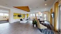 Located at 1040 Jamison Loop in Kissimmee, Florida, this single-story community clubhouse delivers a dynamic and well-appointed gathering space defined by a cohesive modern aesthetic. Bold yellow accents, clean white walls, and warm wood ceiling details unify the interior, creating an energetic yet welcoming atmosphere throughout.

The open lounge area anchors the space with comfortable seating, including a plush sofa and accent chairs arranged around a flat-screen TV, with direct access to an outdoor patio through large glass doors that invite abundant natural light and outdoor views. Adjacent to the lounge, a sleek kitchenette features a long white countertop bar with barstool seating, a wood-paneled tray ceiling, and white subway tile detailing — ideal for social gatherings.

A striking geometric feature wall in yellow, grey, and natural wood tones serves as a bold focal point near the entry, accompanied by high-top workstation seating. The game room area offers entertaining options including a pinball machine, an arcade cabinet, a pool table, and a dedicated card or gaming table framed by floor-to-ceiling windows dressed in yellow curtains. Tile and patterned carpet flooring transition fluidly between zones, reinforcing the space's purposeful, modern design throughout.