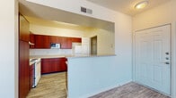 This single-story residence offers a well-organized layout with a cohesive design throughout. The living area serves as the central gathering space, featuring wood-look plank flooring, a ceiling fan, and large windows fitted with blinds that allow natural light to filter in while maintaining privacy. The entry door opens directly into this open, airy space.

The kitchen is positioned adjacent to the living area and is accessible via a pass-through opening, creating an open-concept feel. It features rich, warm-toned cabinetry in a deep reddish-brown finish, light-colored countertops, and white appliances including a refrigerator, electric range, and built-in microwave. Wood-look flooring continues throughout the kitchen, and a utility area with an electrical panel is accessible from within. A peninsula-style half-wall separates the kitchen from the living space.

The bedrooms are carpeted and generously sized, each equipped with a ceiling fan and windows that bring in natural light. At least one bedroom includes access to a walk-in closet with built-in shelving. The bathroom features a single-sink vanity with rich wood cabinetry, a large mirror, a tub-shower combination, and matching wood-look flooring.