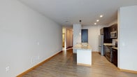 This single-floor space showcases a thoughtfully designed open-concept layout that seamlessly connects the living and kitchen areas. The kitchen is a standout feature, centered around a granite-topped island with an integrated sink and a sleek pendant light overhead. Dark-toned cabinetry pairs elegantly with stainless steel appliances, including a full-size refrigerator, creating a polished and functional culinary space. Recessed lighting illuminates the kitchen area, complementing the neutral white walls and warm hardwood-style flooring that flows consistently throughout the space. The open living area is expansive and bright, offering a clean canvas with ample room for a variety of furniture arrangements. A hallway leads to additional rooms, suggesting a well-organized floor plan with clear separation between living and private areas. Visible floor vents indicate a central HVAC system for year-round climate comfort. The overall aesthetic is contemporary and minimalist, with a neutral color palette that creates a calm, inviting ambiance. The space is well-suited for those who appreciate modern design, open living, and functional, detail-oriented finishes.