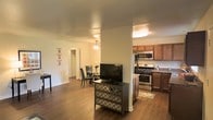 This single-floor residence offers a thoughtfully designed open-concept layout that seamlessly connects the main living areas. The living space is anchored by rich hardwood-style flooring that flows throughout, creating a warm and cohesive aesthetic. A central support column adds architectural character while subtly defining the transition between the living and kitchen areas.

The kitchen is well-equipped with stainless steel appliances, including a gas range, microwave, and refrigerator, complemented by warm wood cabinetry and ample counter space. A decorative area rug adds a welcoming touch near the kitchen entry.

Adjacent to the living area, the dining space features a ceiling fan for added comfort and is furnished with a round table and chairs, ideal for casual meals. A flat-screen television rests atop an ornate mirrored dresser, adding a stylish focal point to the living space.

Neutral-toned walls throughout contribute to a bright and airy ambiance, while the open sightlines between rooms enhance the sense of spaciousness. A hallway visible from the main living area leads to additional rooms, suggesting a practical and functional floor plan. Overall, the space combines comfort and style in a compact, efficient layout.
