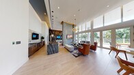 This expansive single-floor space showcases an open-plan layout with soaring double-height ceilings that create an immediate sense of grandeur. The architectural design centers around a dramatic floor-to-ceiling concrete fireplace wall, accented with gold detailing, which serves as the room's focal point and anchors the main living area. Light wood-toned tile flooring runs throughout, lending warmth and continuity across the space.

A wall of floor-to-ceiling windows and glass doors along one side floods the interior with natural light while offering views of lush palm trees and the surrounding outdoor terrace. This seamless indoor-outdoor connection enhances the bright, airy ambiance throughout.

The social hub of the space features a long, dramatic bar counter finished in bold, veined black marble, complemented by warm wood cabinetry and pendant globe lighting. Behind it, a full-height built-in bookcase with integrated lighting adds depth and a sophisticated library aesthetic.

Multiple seating arrangements are thoughtfully positioned throughout — including a lounge area with contemporary rust-toned armchairs, a neutral sofa grouping anchored by a patterned area rug, and a casual dining area with natural wood chairs. A secondary lounge nook with a plush grey sofa and sculptural brass accent tables completes the layout. The overall ambiance is modern, warm, and inviting.