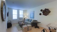 This single-floor living space offers a clean, contemporary design characterized by a neutral color palette of soft whites and warm beiges. The living area is well-lit by two large windows that allow abundant natural light to fill the room, creating an airy and welcoming atmosphere. Light hardwood flooring runs throughout, adding warmth and continuity to the space.

The living area is thoughtfully arranged with a plush gray sectional sofa accented by light blue throw pillows, complemented by a classic wingback chair in a neutral tone. A dark wood dining or console table anchors one side of the room, while textured stone-stacked cube ottomans serve as both functional and decorative accents. A patterned area rug defines the seating zone, adding visual depth.

Wall art plays a notable role in the room's character — a large blue abstract canvas painting adds a pop of color, while dark sculptural floral wall pieces provide an artistic focal point. The overall ambiance is calm, modern, and curated, reflecting a balance between comfort and refined aesthetic sensibility.