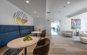 This thoughtfully designed single-floor amenity space offers a sophisticated blend of social, recreational, and professional environments, all unified by a contemporary aesthetic. The main lounge area is anchored by bold blue accent chairs arranged around a sleek fireplace, with textured wall art and vibrant abstract paintings adding artistic character to the crisp white walls. Light-toned, patterned carpet flooring flows throughout the communal zones, creating a warm yet refined atmosphere.

A dedicated social lounge features a built-in navy banquette with café-style seating alongside a neon wall installation, ideal for informal gatherings. A separate sitting room with cream upholstered chairs and floor-to-ceiling glass doors opens toward an outdoor terrace, bathing the space in generous natural light and providing views of the surrounding neighborhood.

The games and entertainment area includes a pool table and a modular blue sectional sofa, complemented by dark hardwood flooring and large windows that connect the interior to the outdoors. A well-appointed kitchenette offers granite countertops, dark cabinetry, stainless steel appliances, a textured wave-tile backsplash, and bar seating.

A fully enclosed glass-walled conference room provides a professional workspace with a central conference table and task seating, seamlessly integrated within the open-plan layout without disrupting the overall flow of the space.