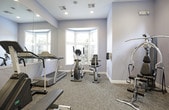 This fitness center offers a comprehensive workout environment designed to support a range of exercise routines. The space is finished with soft blue-gray walls, neutral carpet flooring, and recessed lighting, creating a clean and focused atmosphere throughout.

The cardio area is anchored by a large wall-to-wall mirror that visually expands the room, and features a Matrix treadmill, an elliptical trainer, and a stair climber. Multiple windows along this section allow natural light to filter in, providing views of surrounding greenery.

A dedicated strength training zone includes a multi-function cable and resistance machine positioned near floor-to-ceiling windows that further brighten the space. A two-tier dumbbell rack holds a full range of free weights, accompanied by a flat weight bench for versatile training options.

A wall-mounted flat-screen television and a wall clock are positioned near the entrance, adding convenience for tracking workouts. A water cooler is also available. The fitness center is accessed through a glass-paneled door that opens to an outdoor area, reinforcing the connection to the natural surroundings and contributing to an open, airy feel.