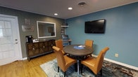 This single-floor space is designed to provide a functional and professional environment well-suited for meetings, consultations, or small group gatherings. The interior is defined by teal-blue accent walls that lend a calm yet polished atmosphere, complemented by warm hardwood flooring and white crown molding that frames the space cleanly.

At the heart of the room sits a round dark-wood conference table surrounded by tan leather chairs, creating an intimate setting ideal for collaborative discussions. A wall-mounted flat-screen television is positioned prominently, making it practical for presentations or media sharing. Recessed ceiling lighting provides consistent, even illumination throughout.

Along one wall, a decorative wood credenza offers additional surface space and storage, accompanied by a coffee and beverage station — a thoughtful touch that adds comfort and convenience for guests and clients. A large framed mirror above the credenza enhances the sense of openness within the room. A literature or display rack nearby keeps informational materials organized and accessible.

An area rug in muted tones anchors the seating arrangement and adds a layer of texture to the space. Overall, the room balances a professional aesthetic with warm, welcoming design elements, making it an effective and comfortable space for client-facing or team-oriented activities.