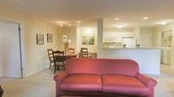 This single-floor residence features an open-concept layout that seamlessly connects the living, dining, and kitchen areas into one cohesive and functional space. The living area is anchored by a warm-toned upholstered sofa, positioned to face the room's interior, creating a welcoming focal point. Neutral-colored carpeting and beige walls contribute to the overall sense of warmth and comfort throughout.

Adjacent to the living space, the dining area accommodates a round wooden table surrounded by classic ladder-back chairs. A floral centerpiece adds a touch of color and life to the setting. Framed artwork on the surrounding walls enhances the space with subtle decorative character.

The kitchen is situated toward the rear and is partially open to the main living area via a pass-through counter, promoting connectivity between spaces. White cabinetry lines the upper walls, complemented by a white refrigerator and microwave, giving the kitchen a clean and classic look. Recessed and flush-mount ceiling lighting provides consistent illumination throughout, ensuring the space remains bright and functional despite limited natural light.