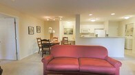 This single-floor residence features an open-concept layout that seamlessly connects the living, dining, and kitchen areas into one cohesive and functional space. The living area is anchored by a warm-toned upholstered sofa, positioned to face the room's interior, creating a welcoming focal point. Neutral-colored carpeting and beige walls contribute to the overall sense of warmth and comfort throughout.

Adjacent to the living space, the dining area accommodates a round wooden table surrounded by classic ladder-back chairs. A floral centerpiece adds a touch of color and life to the setting. Framed artwork on the surrounding walls enhances the space with subtle decorative character.

The kitchen is situated toward the rear and is partially open to the main living area via a pass-through counter, promoting connectivity between spaces. White cabinetry lines the upper walls, complemented by a white refrigerator and microwave, giving the kitchen a clean and classic look. Recessed and flush-mount ceiling lighting provides consistent illumination throughout, ensuring the space remains bright and functional despite limited natural light.