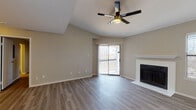 This single-floor residence offers a thoughtfully designed layout with a clean, modern aesthetic evident throughout. The living room serves as the centerpiece of the home, boasting vaulted ceilings that lend a sense of openness and volume to the space. A ceiling fan with integrated lighting adds both style and function, while a white-surround fireplace provides a classic focal point. A sliding glass door floods the room with natural light and provides direct access to an outdoor balcony, offering views of the surrounding landscape. Wood-look flooring runs seamlessly throughout the home, creating a cohesive and contemporary feel. The kitchen is well-equipped with white cabinetry, dark countertops, a subway tile backsplash, stainless steel appliances including a refrigerator and dishwasher, and a single-basin sink. A pass-through window connects the kitchen to the adjacent living area, enhancing the open flow of the floor plan. The bedrooms are comfortably sized, each featuring ceiling fans and the same wood-look flooring. One bedroom includes convenient en-suite bathroom access. The bathroom is finished with a floating wood-toned vanity, a large mirror with vanity lighting, a white countertop, and a shower-tub combination, presenting a clean and modern finish throughout.