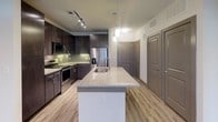 Located at Aura in Grand Prairie, Texas, this single-story unit presents a contemporary design with cohesive finishes throughout. The kitchen serves as a focal point, showcasing an oversized island topped with polished granite countertops and an undermount sink. Dark espresso cabinetry extends to the ceiling, complemented by a green glass tile backsplash and a full suite of stainless steel appliances, including a French door refrigerator and electric range. Pendant lighting and track lighting illuminate the space effectively. Light wood-style flooring runs through the main living and kitchen areas, creating a warm, consistent aesthetic.

The living area is open and well-lit, benefiting from large windows fitted with roller shades that allow diffused natural light to fill the room. A ceiling fan adds functionality to the space.

The bedroom features plush carpeting, a window with blinds, and a ceiling fan, offering a comfortable and quiet retreat. An adjoining walk-in closet provides generous storage with multiple hanging rods and shelving.

The bathroom is well-appointed with a dual-sink vanity topped with granite, dark cabinetry with brushed nickel hardware, a soaking tub, and large-format tile surround. Wood-style flooring continues here, tying the design together cohesively.