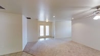This single-story space presents a clean, open layout characterized by a neutral color palette throughout. The main living area is finished with light beige carpeting, lending a warm and understated feel to the room. Recessed lighting and flush-mount ceiling fixtures illuminate the interior evenly, complementing the smooth, textured ceiling overhead. A central entryway features double doors with glass panels that allow natural light to filter into the space, creating a welcoming transition from exterior to interior. The entry area transitions seamlessly from tile flooring to the carpeted living space, providing a practical and visually defined boundary. The walls are painted in a crisp, bright white, contributing to an airy and open ambiance. A ceiling vent is visible near the entryway, indicating a central air system. The overall design is minimalist and functional, with clean lines and an absence of decorative detail, making the space highly adaptable to a variety of interior arrangements and personal styles. The open floor plan allows for flexible furniture placement and a sense of spaciousness throughout the main living area.