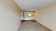 This single-floor unit offers a straightforward, functional layout with clearly defined living and kitchen zones. The open-concept living area is finished with neutral-toned carpet, warm beige walls, and a textured ceiling, creating a simple and clean atmosphere. A ceiling-mounted light fixture provides overhead illumination throughout the main space.

The kitchen occupies a compact footprint at one end of the main living area, transitioning from carpet to hardwood-style flooring. It is equipped with white cabinetry extending to the upper walls for ample storage, a coil-burner electric range, a refrigerator, a single-basin stainless steel sink, and light-colored countertops. An under-cabinet range hood provides ventilation above the stove.

The bedroom is a generously sized, carpeted room featuring a large window dressed with vertical blinds that allows natural light to fill the space. A wall-mounted air conditioning unit and baseboard heating ensure year-round climate comfort.

The bathroom is clean and utilitarian, featuring a full-size soaking tub with a shower rod overhead, a toilet, and textured beige walls that maintain the unit's consistent neutral palette throughout.