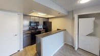 This single-floor unit offers a practical and comfortable layout designed to maximize livability. The open-concept living and dining area features neutral-toned carpet, textured ceilings, and a sliding glass door that floods the space with natural light while providing access to an outdoor area with surrounding views. The transition from carpet to wood-look flooring marks the entrance to the kitchen, which is equipped with dark charcoal cabinetry, granite-style countertops, black appliances including a range, microwave, and refrigerator, and a peninsula that serves as a natural divider between the kitchen and living space.

The bedrooms are generously proportioned, each featuring plush carpeting, neutral wall tones, and windows fitted with vertical blinds that allow for adjustable natural light. The rooms offer a clean, move-in-ready feel with ample wall space for furnishing flexibility.

The bathroom is thoughtfully designed with a long vanity topped with dark granite-style countertops, dark cabinetry with brushed nickel hardware, and a large mirror with overhead lighting. Wood-look flooring extends throughout the bathroom, and a separate tub-shower combination with white tile surround provides a clean, functional bathing area. Overall, the unit presents a cohesive and updated aesthetic throughout.