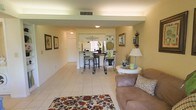 This single-story space showcases an open-concept layout that seamlessly connects the living area, dining nook, and kitchen. The living room is anchored by light beige tile flooring, which runs throughout the space, creating a cohesive and clean aesthetic. A floral area rug adds a pop of color, complementing the warm, neutral-toned walls. The living area features a plush sofa with decorative pillows, a round side table with a table lamp, and framed botanical wall art, contributing to a comfortable and inviting atmosphere. A wrought-iron bistro-style dining set with bar stools occupies the central dining nook, offering a casual yet stylish place for meals. The kitchen sits just beyond, separated by a partial wall with a pass-through counter, allowing natural light and visibility to flow between spaces. Wall-mounted sconces and ceiling-mounted light fixtures provide consistent, soft illumination throughout. Decorative accents, including a wall-mounted candle sconce, potted greenery, and a decorative plate, add character and warmth to the overall design. The layout is efficient and well-organized, making practical use of the available space.