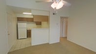 This single-floor space presents an open and functional layout that connects the kitchen and living area seamlessly. The kitchen is defined by warm wood-toned cabinetry, a white refrigerator, and tile flooring, with a half-wall peninsula that serves as a natural divider between the cooking and living spaces. Under-cabinet lighting provides focused illumination over the work surfaces. The living area features neutral wall-to-wall carpeting, contributing to a softer, quieter atmosphere. A ceiling fan with an integrated light fixture is centrally positioned, providing both ventilation and overhead lighting throughout the open space. The transition between the tiled kitchen floor and the carpeted living area creates a clear functional boundary without the use of walls. A hallway visible from the main living space leads to additional rooms, suggesting a straightforward and efficient floor plan. The walls throughout are painted in a clean, neutral off-white tone, keeping the interior feeling bright and versatile. The overall ambiance is practical and understated, with an emphasis on usability and ease of movement throughout the space.
