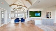 This single-story clubhouse is a well-appointed community amenity space designed with a coastal-contemporary aesthetic. The main entry and leasing area immediately impresses with soaring vaulted ceilings accented by exposed wood beams and a statement chandelier. A dramatic living moss feature wall serves as a striking backdrop to the reception desk, which is finished in light wood and stone-textured tile. Adjacent workstations with herringbone-patterned carpeting provide a functional leasing office environment.

The lounge areas are thoughtfully arranged throughout the space, featuring plush navy blue velvet seating, neutral sofas, and natural wood coffee tables. Tall transom windows and glass French doors flood the interiors with natural light while offering outdoor views. A media wall with a mounted flat-screen television and woven pendant lighting anchors one of the main gathering areas.

A dedicated games room includes a full-size billiards table, a wall-mounted cue rack, bar-height counter seating, and direct access to an outdoor deck via a glass door. Warm wood-look flooring runs throughout most of the space, unifying the various zones. The overall ambiance is bright, modern, and inviting, with carefully curated décor and greenery reinforcing the coastal atmosphere.