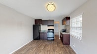 This single-floor unit offers a practical and functional layout with clearly defined living spaces. The kitchen is a standout feature, equipped with dark espresso cabinetry, granite-style countertops, and black appliances including a full-size refrigerator and gas range. A mosaic tile backsplash adds a decorative touch near the sink area, which benefits from natural light through a window fitted with blinds. The kitchen flows seamlessly into an open living area finished with durable wood-look laminate flooring, neutral gray walls, and a textured ceiling with flush-mount lighting, creating a bright and cohesive space. A separate bedroom is finished with plush wall-to-wall carpeting, neutral tones, and a window that allows natural light to filter in, providing a comfortable and quiet retreat. The bathroom is well-appointed with white tile surround, a tub/shower combination, a granite-topped vanity, and a large mirror. Clean white walls and wood-look flooring complete the bathroom's tidy, functional aesthetic. Throughout the unit, consistent neutral color tones and modern finishes contribute to a clean, move-in-ready atmosphere.