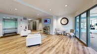 Located at 3030 S Water St in Pittsburgh's South Side neighborhood, this single-floor amenity space offers residents two thoughtfully designed areas: a lobby lounge and a dedicated exercise room. The hallway and lobby area greets visitors with warm, light-toned hardwood flooring, recessed lighting, and a clean, contemporary palette of soft whites and blues. Comfortable seating arrangements — including white upholstered chairs and sofas accented with patterned throw pillows — create a welcoming social environment. A wall-mounted display, decorative clock, and a full bank of resident mailboxes add both function and character to the space. Large glass entry doors allow abundant natural light to pour in, enhancing the open, airy atmosphere. The adjacent exercise room is equally well-appointed, featuring large floor-to-ceiling windows that frame lush greenery and provide ample daylight throughout workouts. Cardio equipment including treadmills, stationary bikes, and a recumbent bike are complemented by a full dumbbell rack, weight bench, and an assortment of fitness accessories such as stability balls and resistance tools. A mirrored accent wall visually expands the room, while durable rubber flooring and a drop ceiling with recessed lighting complete the polished, practical design. Glass doors connect the two spaces seamlessly.