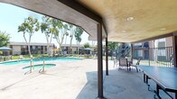 Located at 9091 Holder St in Cypress, California, this outdoor community amenity space offers a well-appointed recreational environment designed for relaxation and social activity. At the heart of the space is a generously sized swimming pool with a curved, inviting shape, complemented by an adjacent spa — both maintained to a high standard and surrounded by a broad concrete pool deck. Lounge chairs and umbrella-shaded seating are arranged poolside, providing ample space for sunbathing and casual gatherings beneath the Southern California sky. Mature eucalyptus trees frame the perimeter, offering natural shade and a lush, park-like atmosphere. A covered patio pavilion with a stucco ceiling and recessed lighting extends the usable outdoor area, sheltering a ping-pong table and additional seating — ideal for leisure and social interaction. An outdoor shower station with tile surround is conveniently situated nearby for post-swim use. Iron fencing defines the boundaries of the pool area while maintaining open sightlines to the surrounding landscaped grounds, which feature neatly maintained grass lawns. The overall setting reflects a resort-inspired design, providing residents with a comfortable and attractive outdoor retreat in a well-maintained community environment.