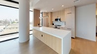 This contemporary single-floor residence showcases a sleek, modern design defined by clean lines, an all-white palette, and abundant natural light. The open living area is anchored by dramatic floor-to-ceiling black-framed windows that wrap around a corner, offering sweeping urban views and flooding the space with daylight. A sculptural white cylindrical column adds an architectural focal point to the main living area, complementing the light-toned wide-plank hardwood flooring found throughout.

The kitchen is thoughtfully designed with flat-front light wood cabinetry extending to the ceiling, stainless steel appliances including a built-in oven, microwave, and gas cooktop, and a generous island topped with a crisp white surface. A linear pendant light above the island reinforces the space's contemporary aesthetic.

The private rooms feature the same light hardwood flooring, bright white walls, and access to natural light through floor-to-ceiling windows. Two bathrooms serve the unit: one features large-format dark tile flooring, a glass-enclosed walk-in shower with marble-style tile surround, and a light wood vanity; the other includes a soaking tub with a frameless glass enclosure and matching marble-style tile surround. The overall ambiance is calm, refined, and distinctly modern.