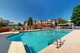 This outdoor amenity space centers around a generously sized swimming pool with crystal-clear turquoise water, offering a resort-style atmosphere designed for relaxation and recreation. The pool features a gently curved design with multiple entry points, including stainless steel handrails, and is finished with a durable aggregate deck surface that surrounds the water on all sides.

The expansive pool deck provides ample space for lounging, with numerous chaise lounge chairs positioned throughout the area for sunbathing. Two modern pergola structures with white canopies offer shaded retreats along the poolside, while additional freestanding patio umbrellas provide flexible shade options near outdoor dining and seating areas. A charming pool house or clubhouse structure sits adjacent to the pool, featuring large windows, a covered porch, and ceiling fans, providing a comfortable indoor-outdoor gathering space.

The entire pool area is enclosed by a white picket fence, offering both security and a clean, classic aesthetic. Mature trees and well-maintained landscaping frame the perimeter, providing natural shade and a lush, park-like setting. The surrounding multi-story brick and siding residential buildings are visible beyond the fence, contributing to a community-oriented ambiance. Under clear blue skies, this outdoor space delivers a bright, welcoming environment ideal for leisure and social activity.