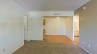This single-floor residence offers a straightforward, functional layout designed to maximize usability. The main living area is finished with soft wall-to-wall carpeting, providing a warm and comfortable foundation underfoot. A textured ceiling adds subtle dimension to the space, while neutral white walls keep the interior feeling bright and clean.

The living area flows openly into an adjacent room — distinguished by hard-surface flooring in a warm tan tone — creating a natural separation between spaces while maintaining an open, connected feel. This secondary space is well-suited for use as a dining area or additional living space and is fitted with a central ceiling light fixture.

A pass-through or partial wall opening connects to a kitchen area visible in the background, which features wood-toned cabinetry that adds a touch of warmth to the overall palette. The kitchen appears to continue along the perimeter, offering practical storage and prep space.

Throughout the residence, a central HVAC vent is integrated into the ceiling, ensuring consistent climate control. A closet with sliding doors is conveniently located near the main living area, providing accessible storage. The overall design is clean, neutral, and move-in ready, offering a versatile canvas suited to a variety of furnishing and décor styles.