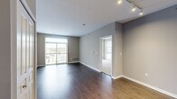 This contemporary single-floor residence offers a thoughtfully designed layout that balances open living spaces with comfortable private areas. The main living area features rich dark hardwood flooring, neutral gray walls, and track lighting overhead, creating a clean and modern aesthetic. A sliding glass door with vertical blinds leads to a private balcony, flooding the space with natural light and offering views of the surrounding area.

The kitchen is a functional standout, outfitted with stainless steel appliances including a refrigerator, electric range, over-the-range microwave, and dishwasher. White cabinetry with warm wood-toned lower cabinets pairs with granite countertops and a peninsula that provides additional prep and dining space. Light hardwood flooring ties the kitchen seamlessly into the living area.

The bedrooms are finished with plush wall-to-wall carpeting in a neutral tone, offering a soft contrast to the hard flooring elsewhere. Multiple windows with horizontal blinds allow for ample natural light while maintaining privacy. The bathroom is cleanly appointed with a white vanity, a natural light-providing window, and classic fixtures. Throughout the unit, neutral taupe and gray tones create a calm, cohesive ambiance well-suited for modern living.