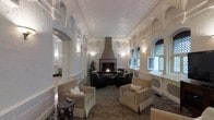 This sophisticated lounge exudes classic architectural grandeur, characterized by its striking vaulted ceiling adorned with elaborate plasterwork and ornamental detailing that immediately draws the eye upward. The space is anchored by a central fireplace fitted with a substantial hood, serving as the room's focal point and surrounded by a carefully arranged seating group of dark upholstered armchairs and a coffee table, ideal for intimate gatherings. Flanking the perimeter, additional seating in neutral tones provides flexible arrangements for guests. The walls feature arched recesses and Gothic-style leaded windows with decorative lattice patterning, allowing soft, diffused natural light to filter into the interior. Wall-mounted sconces supplement the lighting, casting a warm glow that highlights the textured plasterwork throughout. Structural columns with ornate detailing define distinct zones within the elongated room, while a richly patterned area rug grounds the seating area and adds warmth underfoot. The overall atmosphere is one of timeless elegance, blending historic architectural elements with comfortable furnishings to create a welcoming yet stately environment.