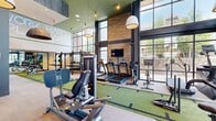 Located at 2101 Bond Street in Charlottesville, VA, this expansive fitness center is designed to deliver a premium workout experience. The single-floor facility spans a generous footprint and is defined by its double-height ceilings and dramatic floor-to-ceiling steel-framed windows that flood the interior with natural light and offer open views of the surrounding neighborhood.

The layout is thoughtfully organized into distinct training zones. A central indoor turf track runs the length of the space, providing a dedicated lane for functional training, sled pushes, and agility drills. Along one side, a row of True treadmills and cardio machines lines the window wall, allowing users to exercise with abundant natural light. The strength training area features a multi-station power rack system loaded with bumper plates, a full dumbbell rack, and an assortment of cable and selectorized machines. Additional equipment includes stationary bikes and BOSU balance trainers.

Warm wood accent walls contrast with dark charcoal structural columns and green ceiling details, creating a bold, contemporary aesthetic. Large pendant lights hang from the soaring ceiling, complementing the recessed lighting throughout. A wall of mirrors expands the sense of space, while motivational signage adds an energetic character to the environment. A bank of personal storage lockers near the entrance provides convenient storage for users.