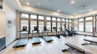 Located at 255 Williamsburg Pkwy in Fate, TX, this single-floor fitness center is thoughtfully designed to offer a comprehensive workout experience in a bright and modern setting. The space is defined by an expansive bank of floor-to-ceiling windows framed in dark trim, flooding the interior with abundant natural light and offering views of the surrounding community, including a glimpse of the outdoor pool area.

The cardio zone lines the windowed wall and features multiple treadmills equipped with individual digital screens, alongside stationary bikes, a stair climber, and elliptical machines — providing a full range of aerobic options. The strength training area is equally well-appointed, with a multi-station cable machine, a weight rack, adjustable benches, and a selection of free weights.

Light wood-style flooring runs throughout, complementing the neutral wall tones of warm beige and soft blue-gray. Large wall mirrors amplify the sense of space and allow users to monitor their form. A flat-screen television is mounted above a built-in storage unit with cubbies and cabinetry, adding a functional and polished finishing touch. Recessed lighting and a ceiling fan complete the comfortable, well-ventilated atmosphere, making this fitness center a standout amenity.