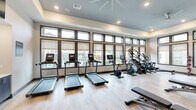 Located at 255 Williamsburg Pkwy in Fate, TX, this single-floor fitness center is thoughtfully designed to offer a comprehensive workout experience in a bright and modern setting. The space is defined by an expansive bank of floor-to-ceiling windows framed in dark trim, flooding the interior with abundant natural light and offering views of the surrounding community, including a glimpse of the outdoor pool area.

The cardio zone lines the windowed wall and features multiple treadmills equipped with individual digital screens, alongside stationary bikes, a stair climber, and elliptical machines — providing a full range of aerobic options. The strength training area is equally well-appointed, with a multi-station cable machine, a weight rack, adjustable benches, and a selection of free weights.

Light wood-style flooring runs throughout, complementing the neutral wall tones of warm beige and soft blue-gray. Large wall mirrors amplify the sense of space and allow users to monitor their form. A flat-screen television is mounted above a built-in storage unit with cubbies and cabinetry, adding a functional and polished finishing touch. Recessed lighting and a ceiling fan complete the comfortable, well-ventilated atmosphere, making this fitness center a standout amenity.