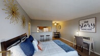 This studio in College Station, Texas offers a thoughtfully organized single-floor layout that maximizes every square foot. The main living area is open and versatile, featuring warm hardwood-style flooring, neutral gray walls, and ample space to accommodate a sleeping area, a dedicated workspace with a desk and chair, and a dining nook with a compact table and stools. Natural light filters in through windows, brightening the space and offering views of the surrounding landscape. A pass-through counter with bar seating connects the living area to the galley-style kitchen, which is equipped with warm wood cabinetry, granite-look countertops, a double-basin stainless steel sink, an electric range, a dishwasher, and a full-size refrigerator — providing a fully functional cooking space within a compact footprint. The bathroom features a wide vanity with a clean white countertop and ample cabinet storage, alongside a separate water closet with a tiled shower-tub combination. Throughout the studio, consistent hardwood-style flooring and a cohesive neutral color palette create a unified, comfortable ambiance well-suited for students or professionals seeking practical, self-contained living in College Station.