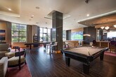 The Club Room at 140 Old Country Rd in Mineola, NY is a thoughtfully designed amenity space that blends modern aesthetics with comfortable, multi-purpose functionality. The single-floor layout is unified by rich dark wood-style tile flooring and deep crimson carpeted zones, creating a dynamic visual contrast throughout the space.

Upon entry through sleek glass-and-black-frame doors, residents are welcomed into an open, airy environment bathed in natural light from expansive floor-to-ceiling windows along the perimeter. A lounge area features a contemporary linear fireplace set against a textured dark accent wall, complemented by plush seating, a sofa, and stylish accent chairs arranged around a marble-top coffee table — ideal for casual gatherings.

A dedicated entertainment zone showcases a large wall-mounted flat-screen TV with bar-height seating, anchored by a curved wood entertainment wall. Crystal chandelier pendant lighting and recessed ceiling fixtures add warmth and sophistication throughout.

The recreation area centers around a full-size billiards table and a shuffleboard table, offering engaging leisure options. A built-in bar counter with high stools provides a social hub, while a bookcase and additional lounge seating near the windows round out the space. Vibrant contemporary artwork adorns the walls, contributing to the room's energetic yet polished ambiance.